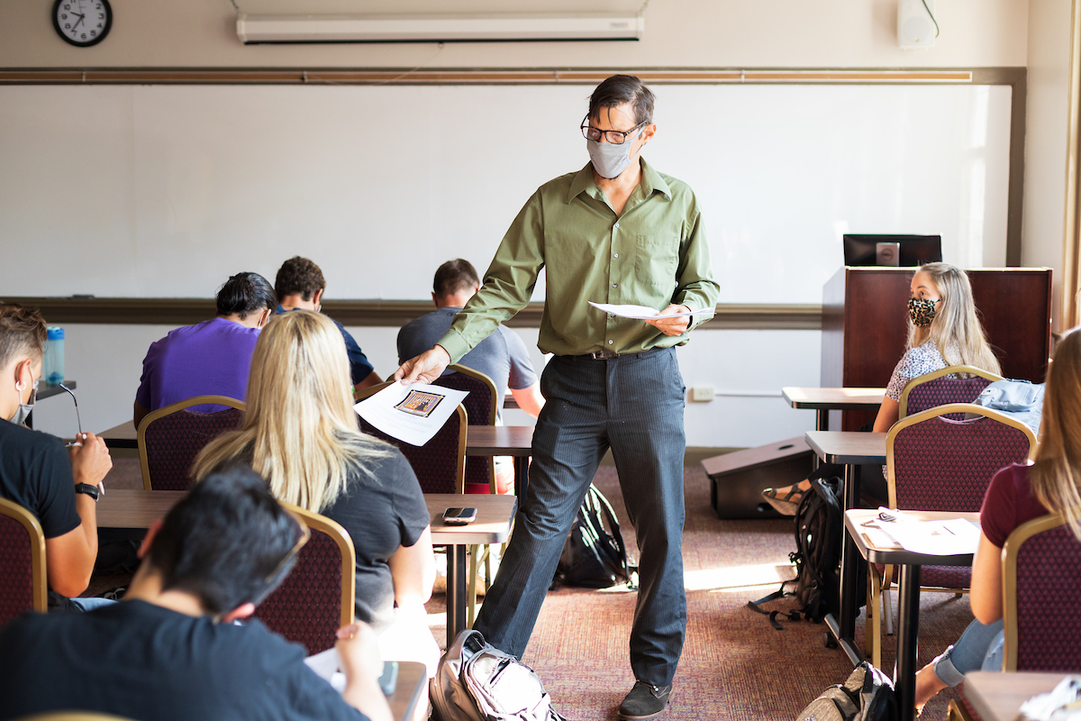 A masked professor hands out papers to masked students on the first day of classes