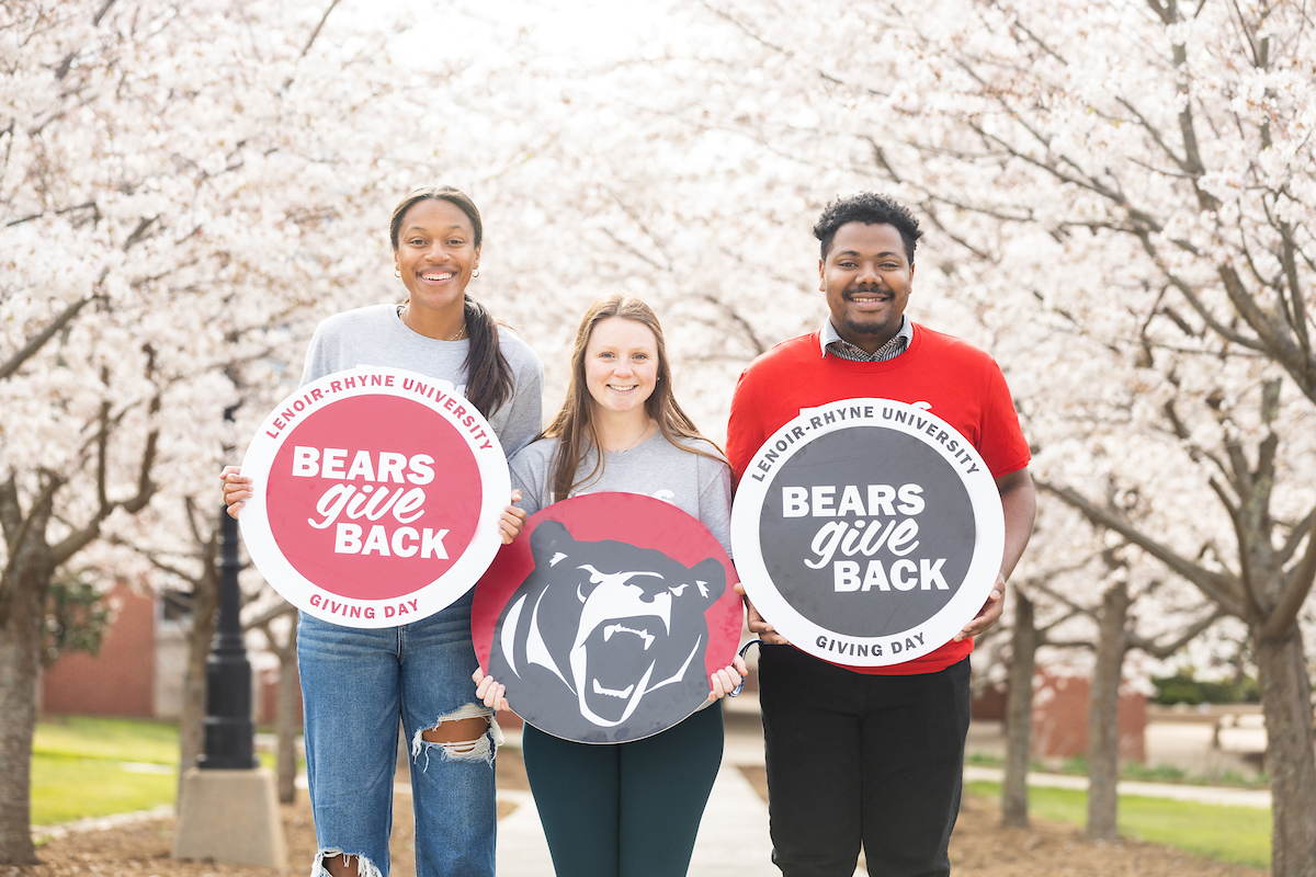 Three students standing outside smile at the camera while holding signs