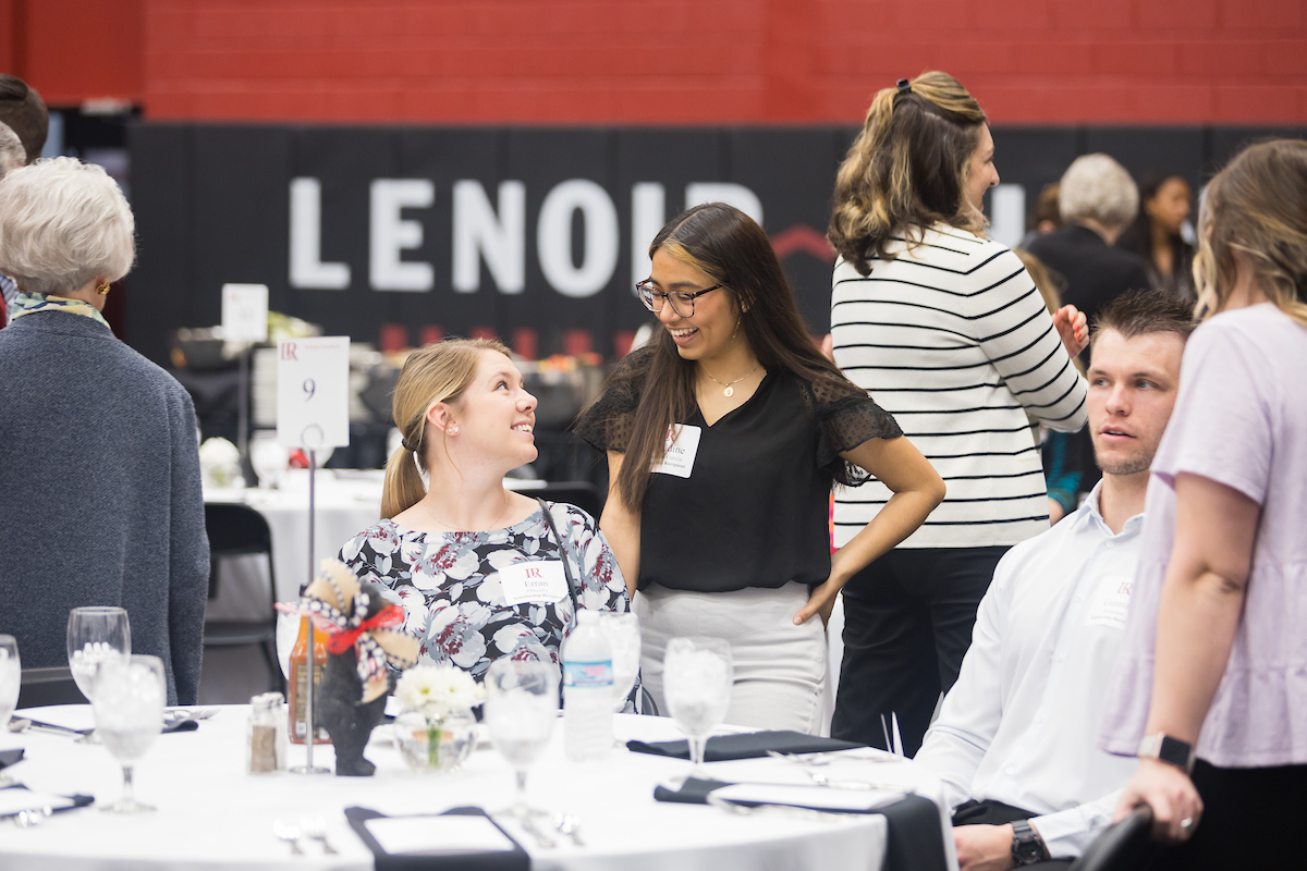 A student sits at a table and speaks to another student standing while inside