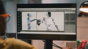 A person sits at a computer that is displaying a robot on the screen