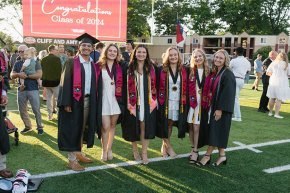 Six students in graduation regalia stand on the field under a sign reading "Congratulations Class of 2024"