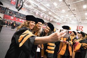 A group of four DNP graduates takes a selfie in their regalia in Shuford Gymnasium 