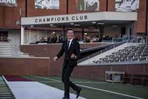 Isaac Riggs wearing a suit and walking across Moretz Field with the club box in the background