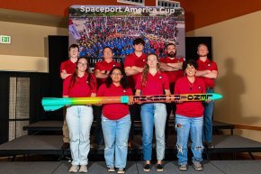 All members of the rocket team stand with their 2024 rocket in front of the Spaceport America Cup logo