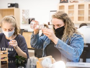 Amy Sain, right, pours liquid from a beaker while masked in a lab