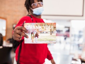 A masked student holds out a thank you postcard to the camera