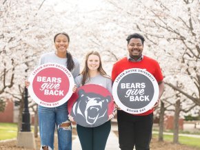 Three students standing outside smile at the camera while holding signs