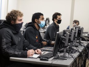 Students sit in a classroom in front of computers while wearing masks