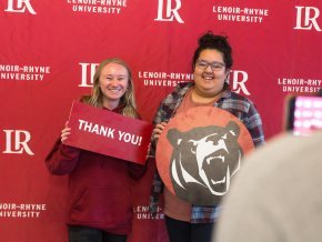 Two students hold up signs against a red and white backdrop