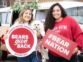 Two female students smile while holding signs and a little dog while outside