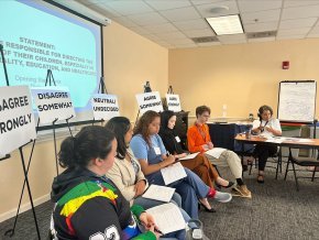 Students sit in a row in front of a screen and a series of placards in a dialogue training session