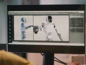 A person sits at a computer that is displaying a robot on the screen