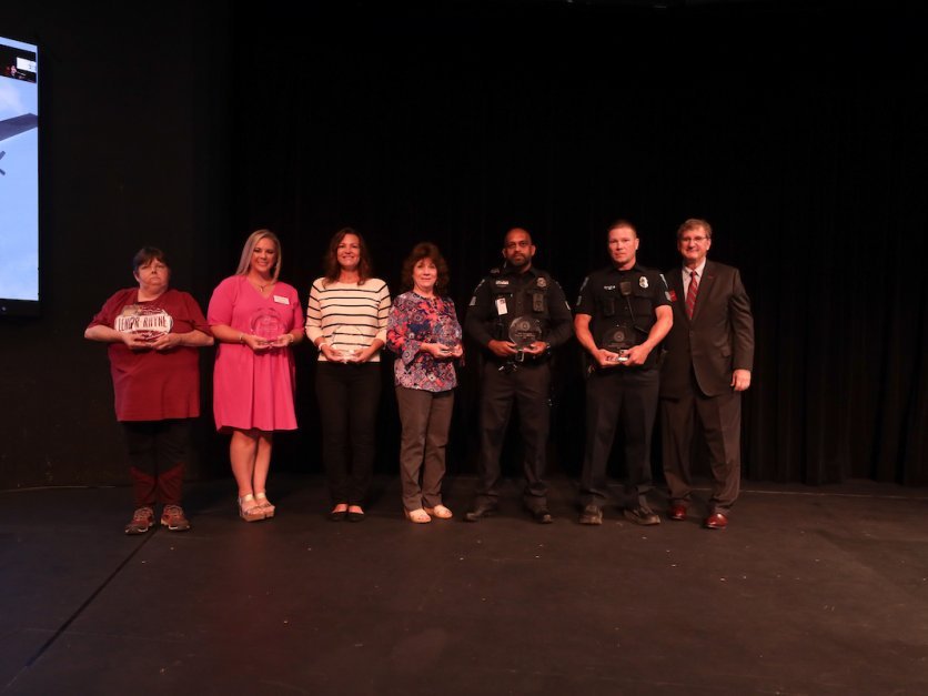 A group of staff members and police offers stand on a stage and smile for the camera indoors