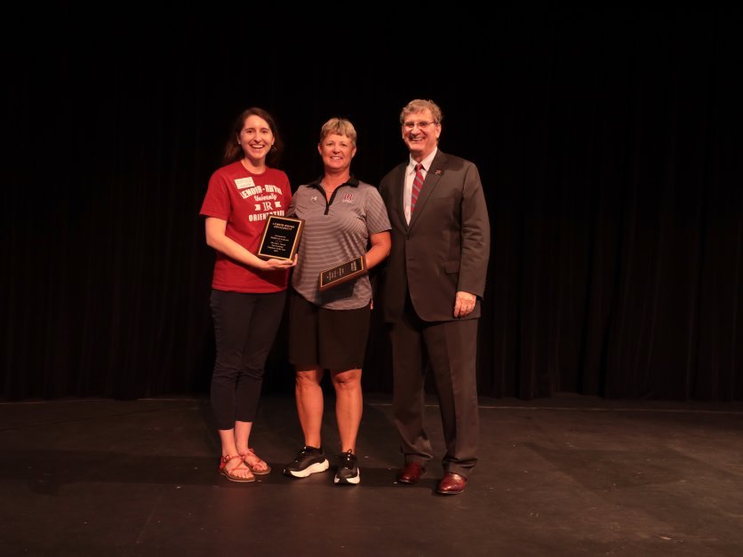 Three people stand on an indoor stage and smile at the camera